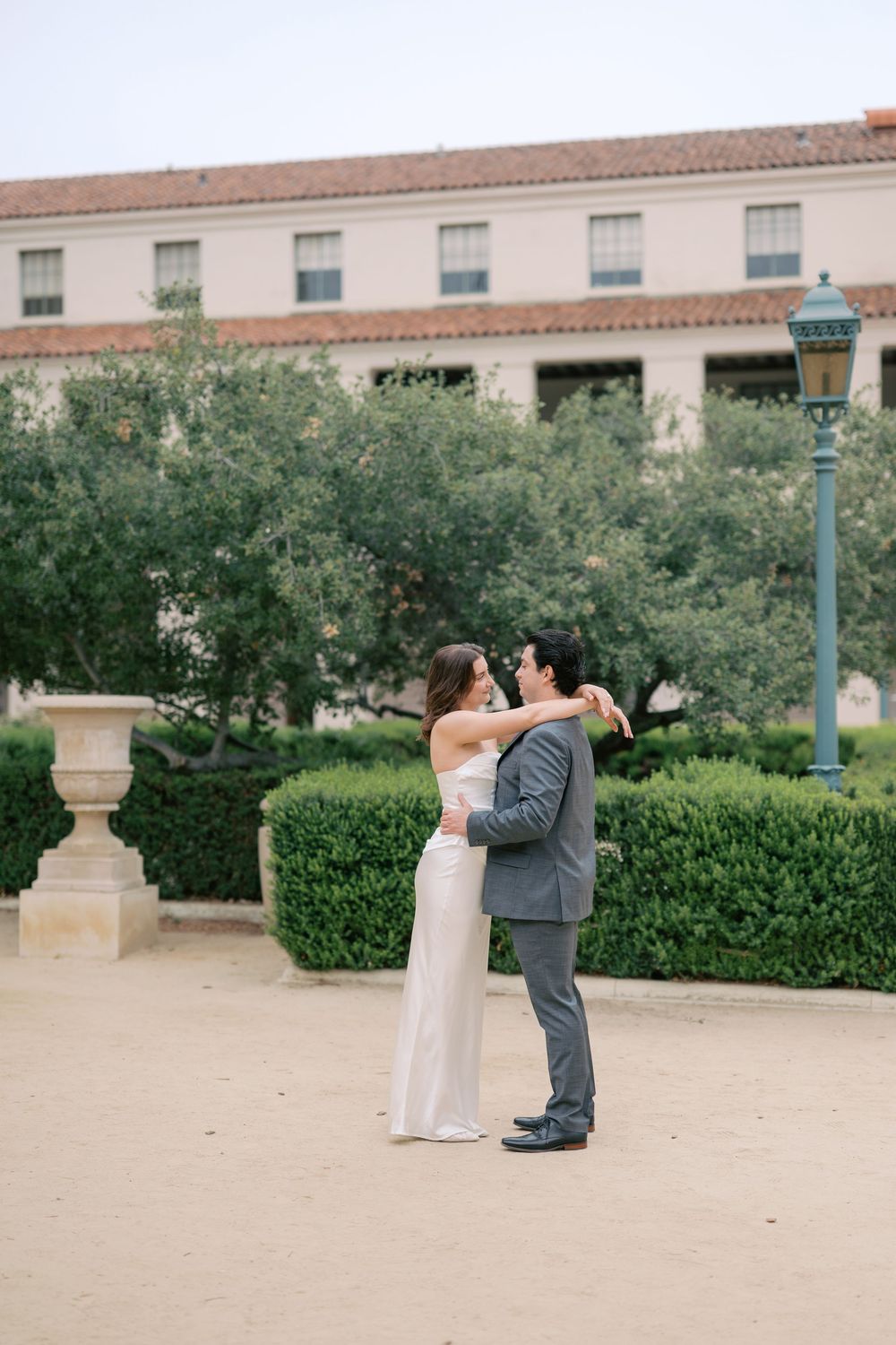 A couple in wedding attire shares a romantic embrace outdoors near manicured hedges and a historic building.