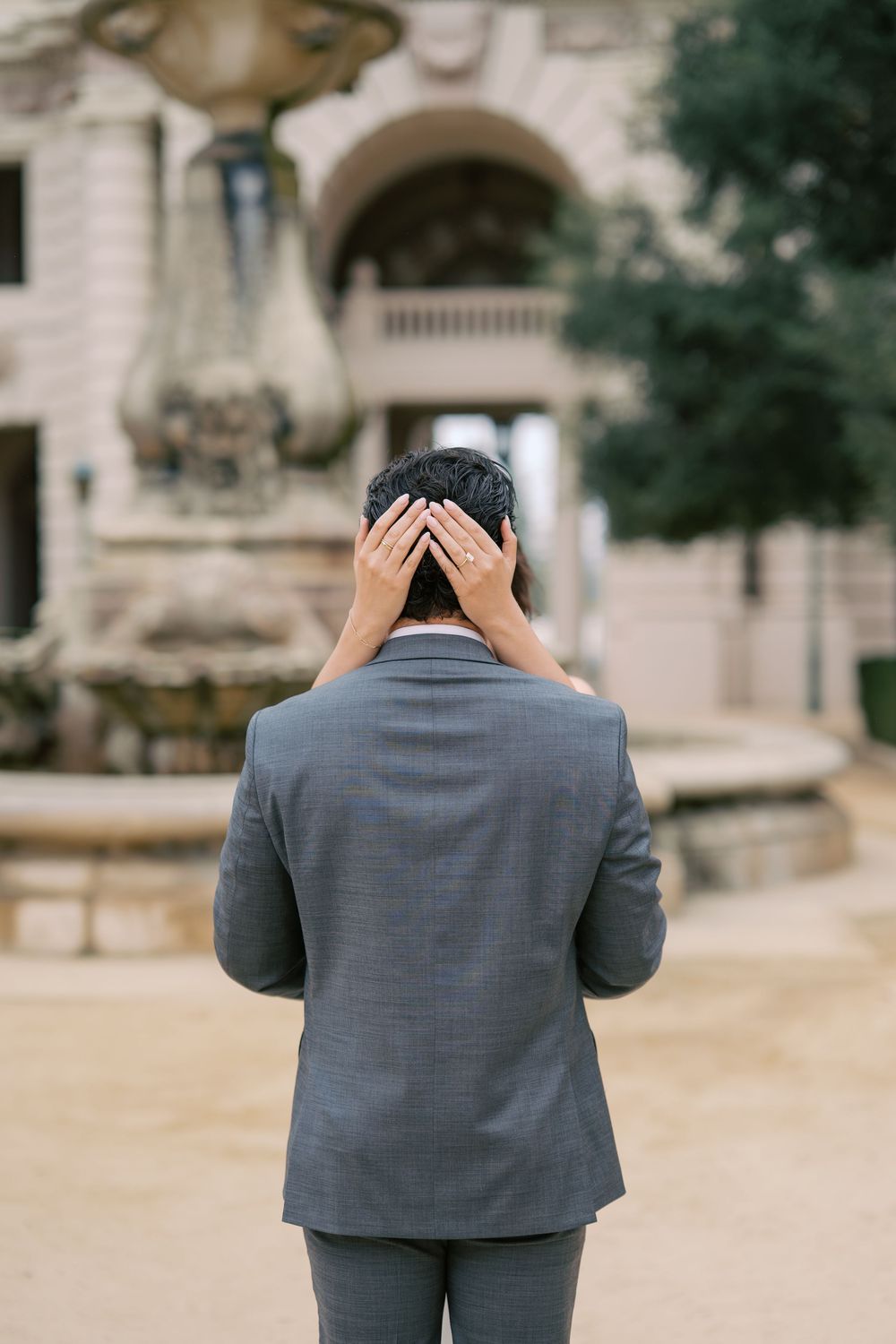 Man in gray suit covering face with hands standing in front of ornate fountain outdoors.
