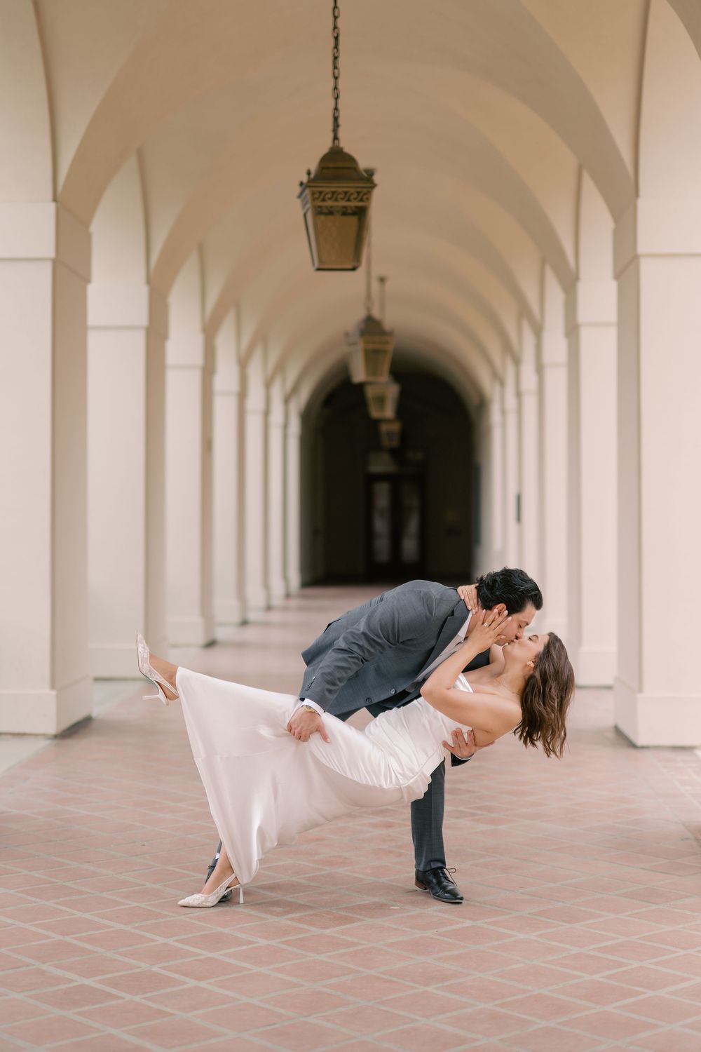 Bride and groom share a romantic dip kiss under arched colonnade corridor during wedding photo session.