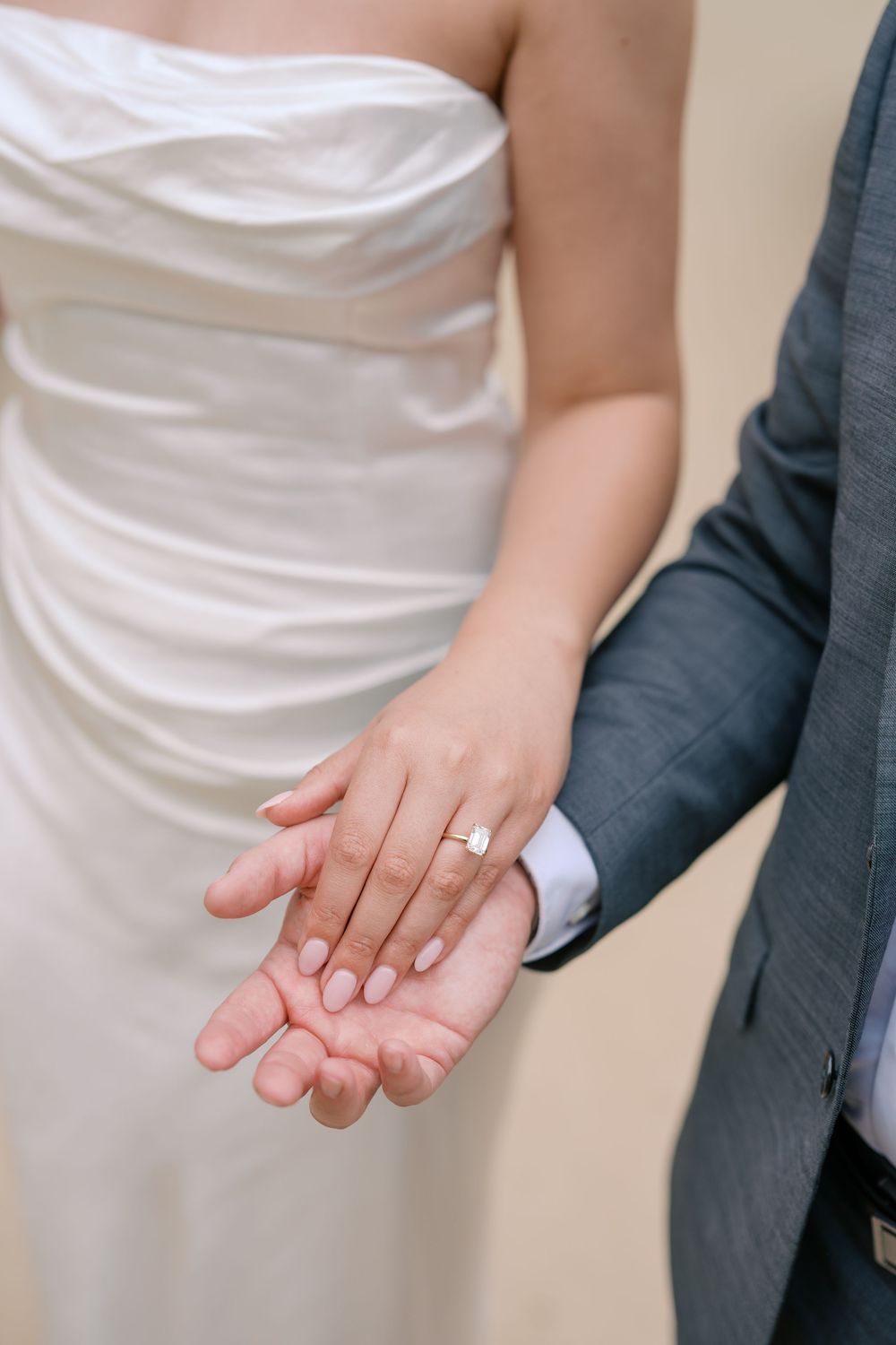 Bride and groom holding hands showing diamond engagement ring, bride in white strapless gown, groom in gray suit.
