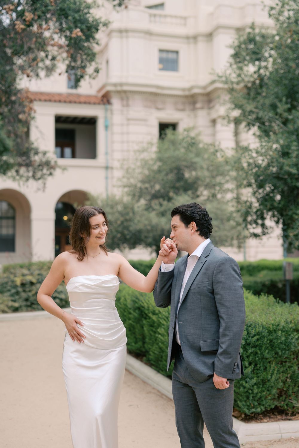 A groom in a gray suit kisses his bride's hand as she smiles in a white strapless gown outdoors.
