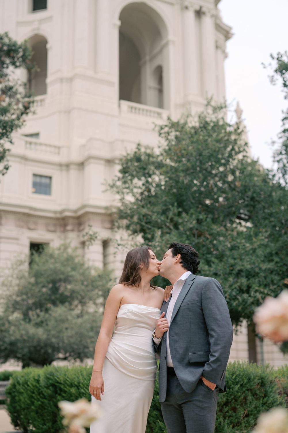 Bride and groom share a romantic kiss outside a grand white building surrounded by lush greenery.