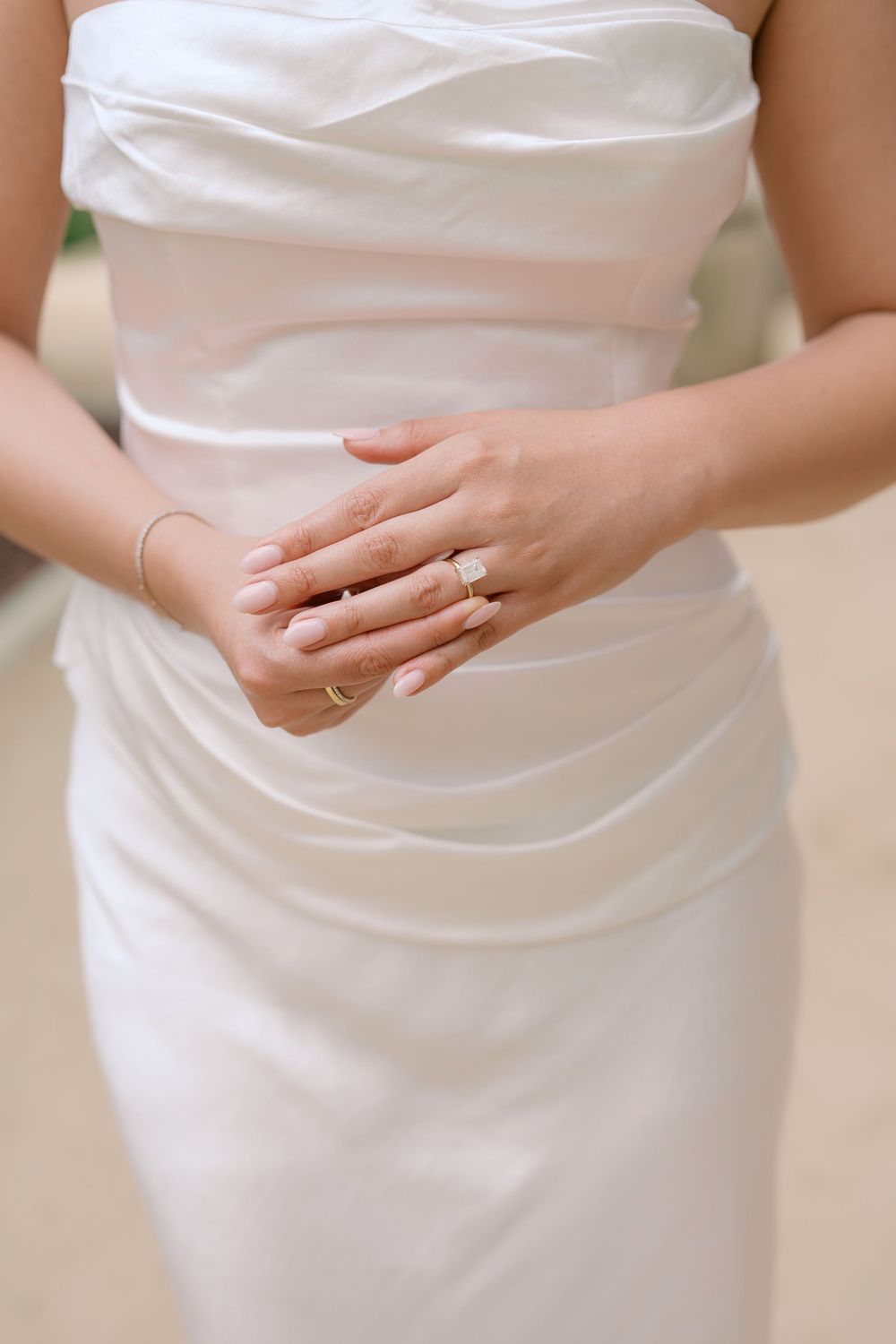 Close-up of bride in strapless white satin wedding gown showing elegant rings and bracelet on clasped hands.