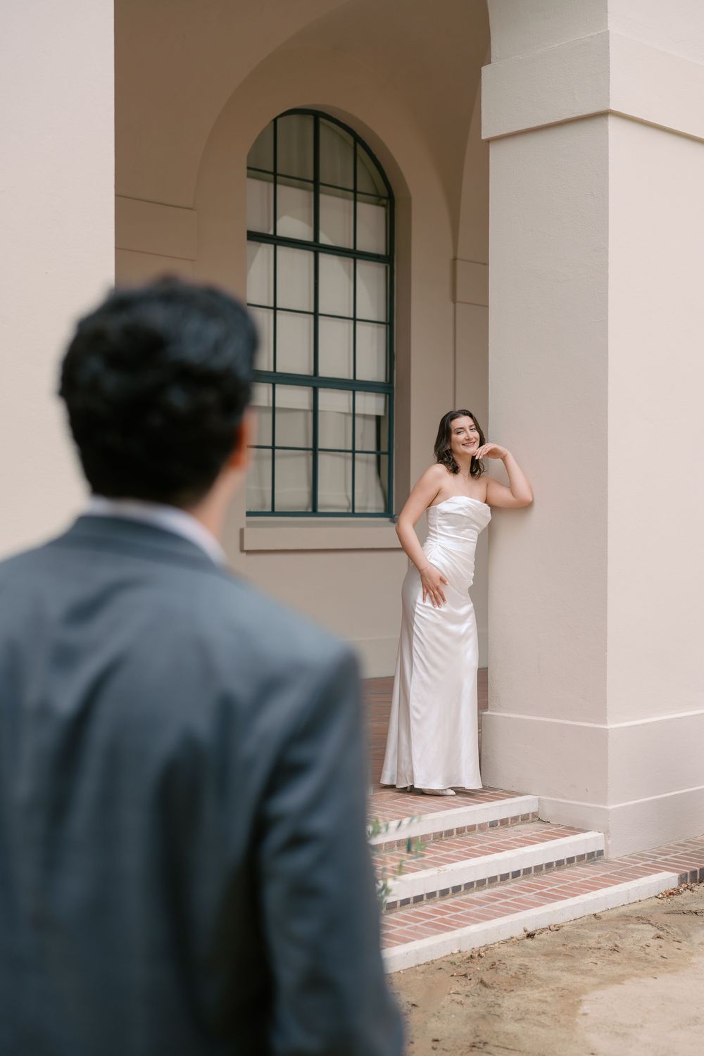 Bride in white satin gown poses by arched window as groom in gray suit approaches during wedding day.
