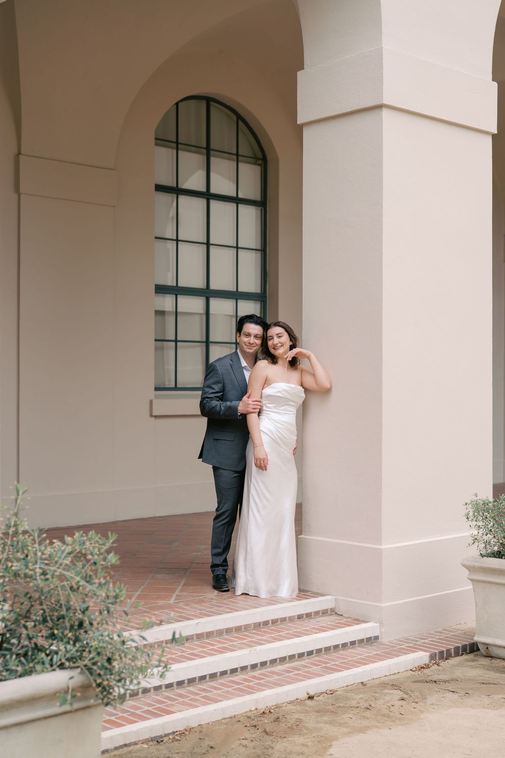 Couple in wedding attire posing on brick steps beside elegant arched columns of a classic building.