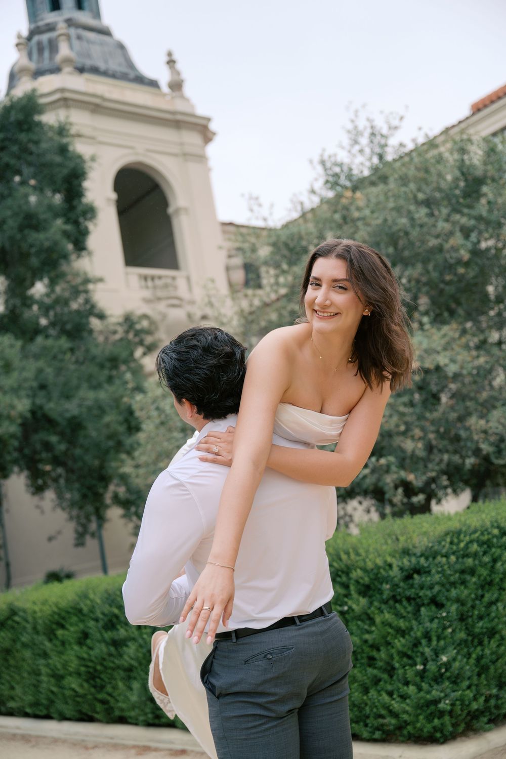 A smiling woman in a strapless top being lifted by a man outdoors near a historic building with lush greenery.