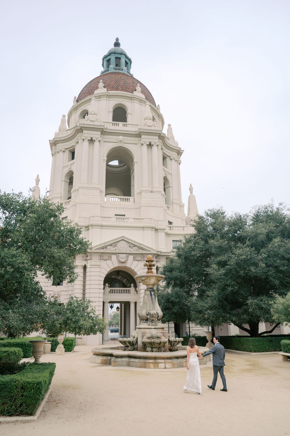 Wedding couple holds hands near ornate fountain outside grand Pasadena City Hall dome building.