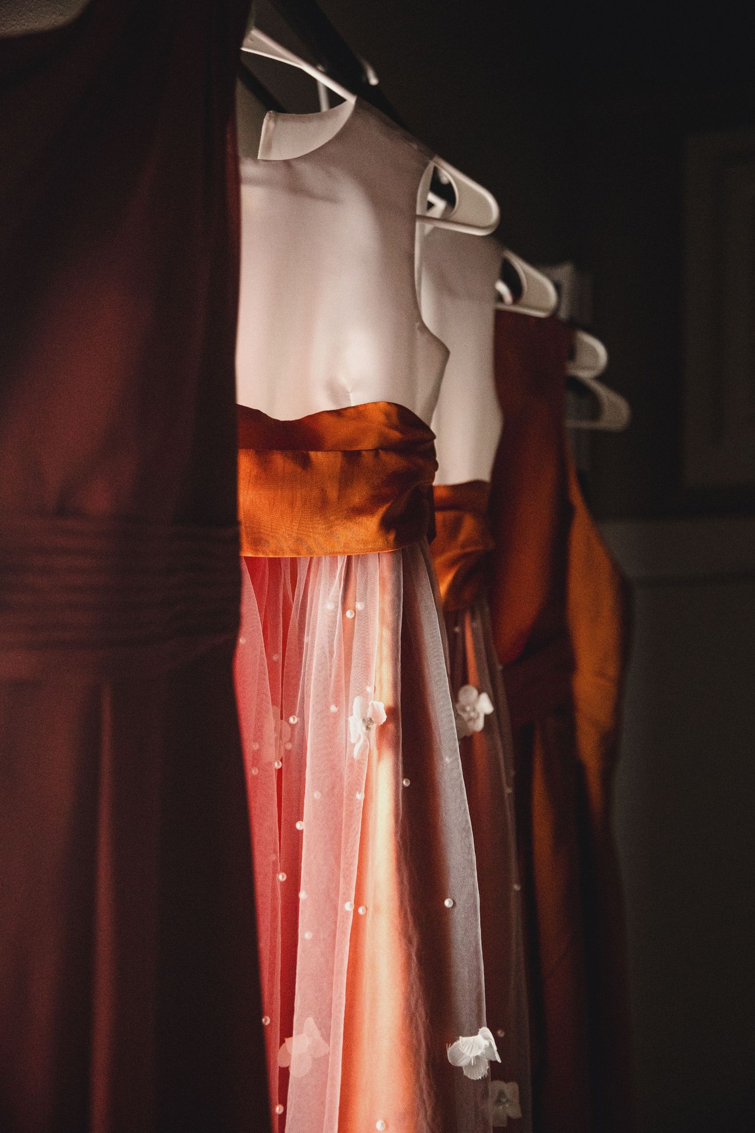 Elegant burnt orange bridesmaid dresses with pearl-embellished tulle overlays hanging on hangers in a dark room.