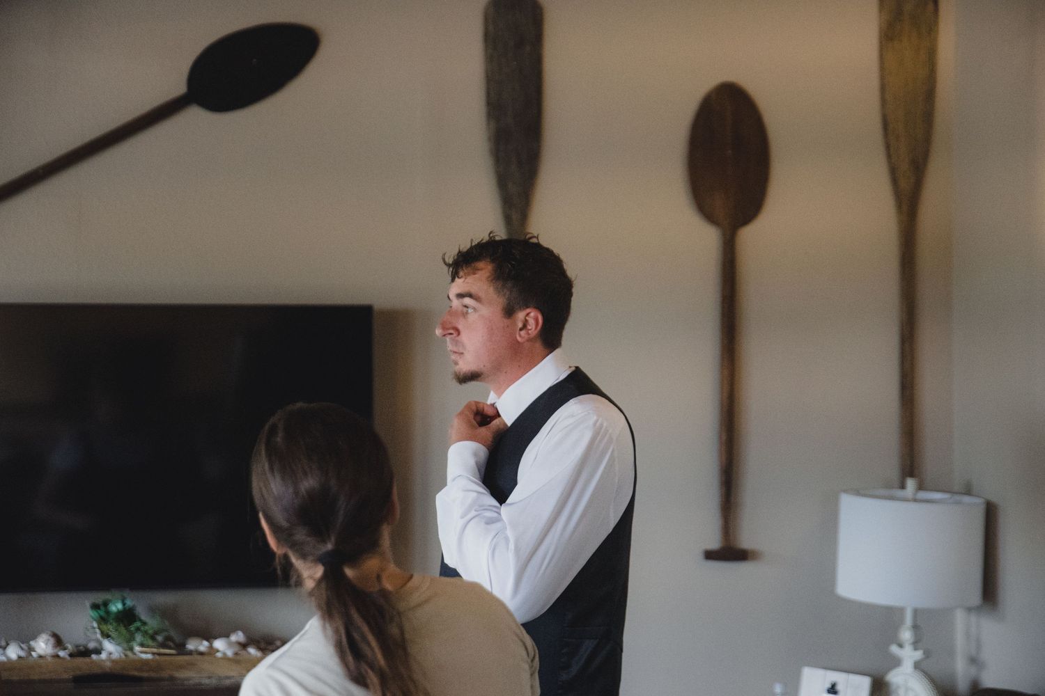 Groom in white shirt and black vest adjusting his tie while getting ready indoors with decorative oars on wall.