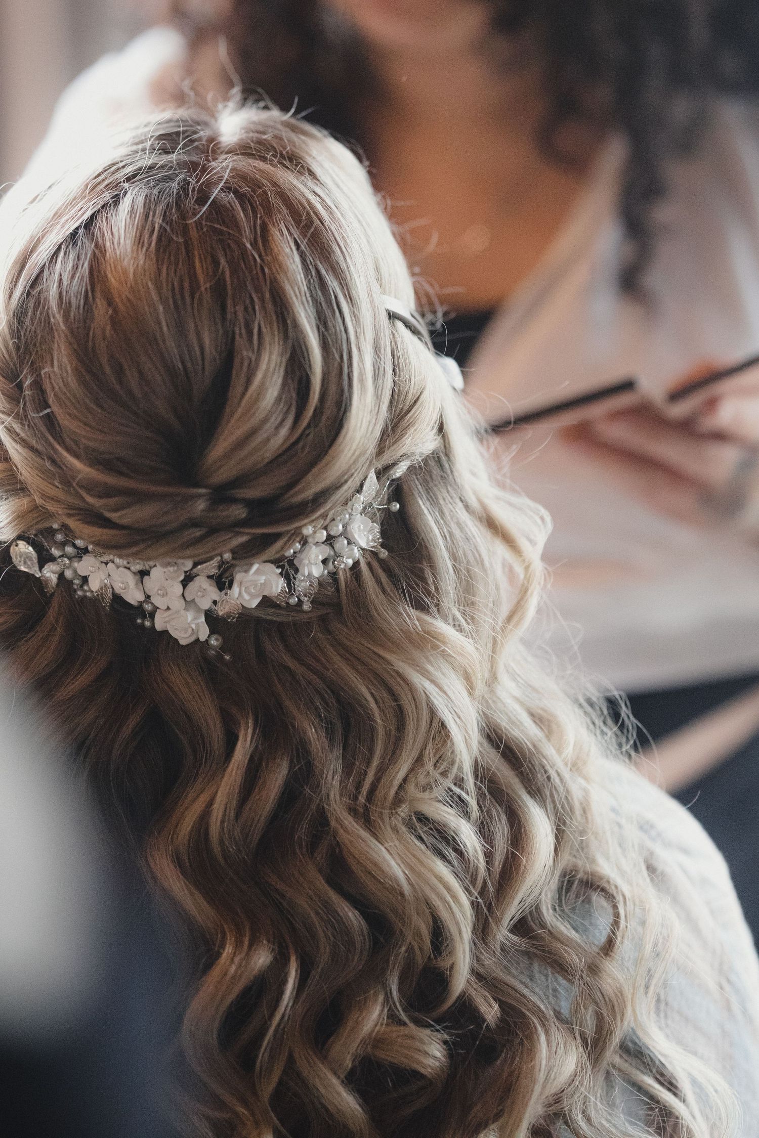 Bride with long curly hair adorned with a delicate floral hair vine, being styled by a hairdresser.