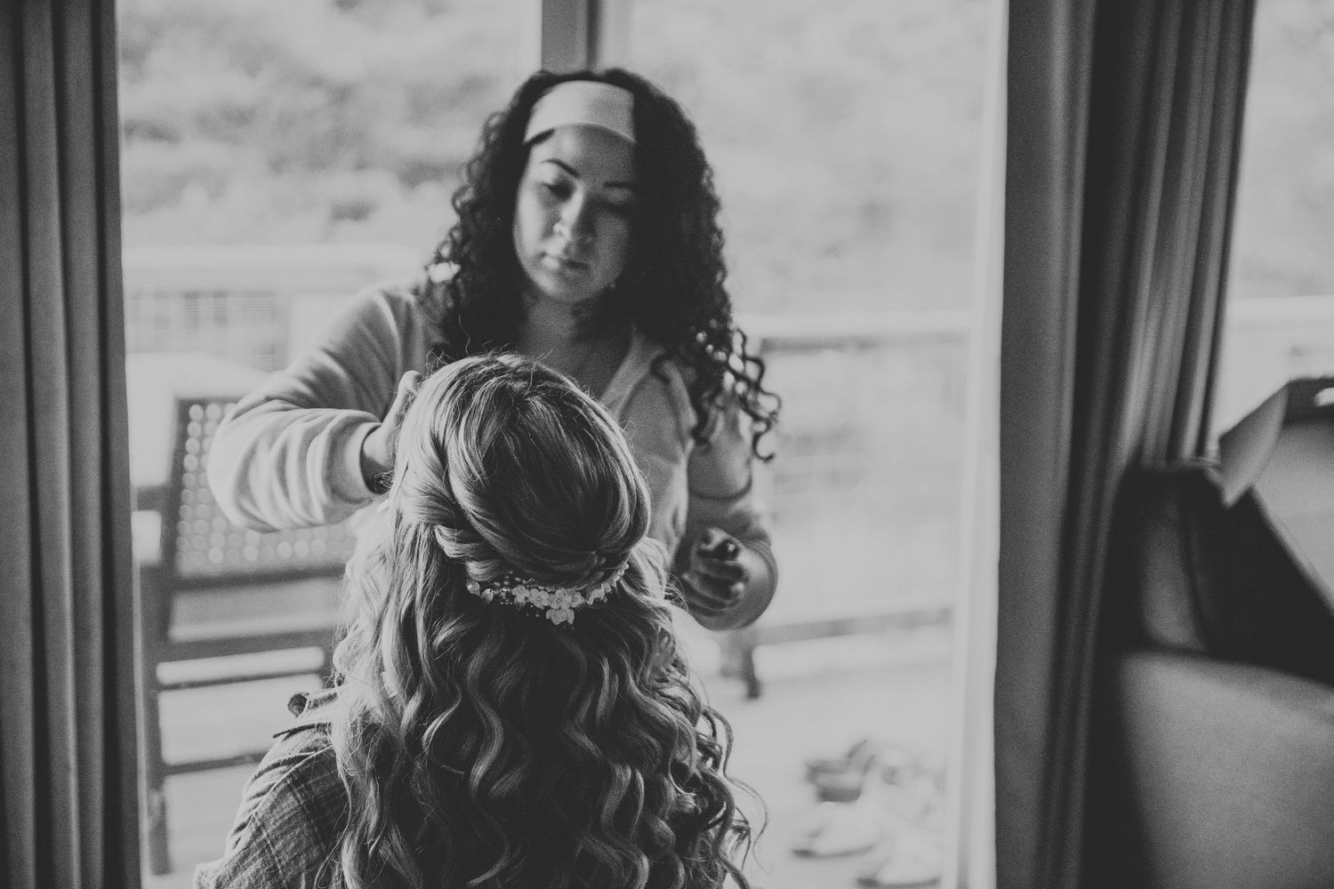 Black and white photo of a hairstylist styling a bride's curly hair with a floral headpiece before her wedding.