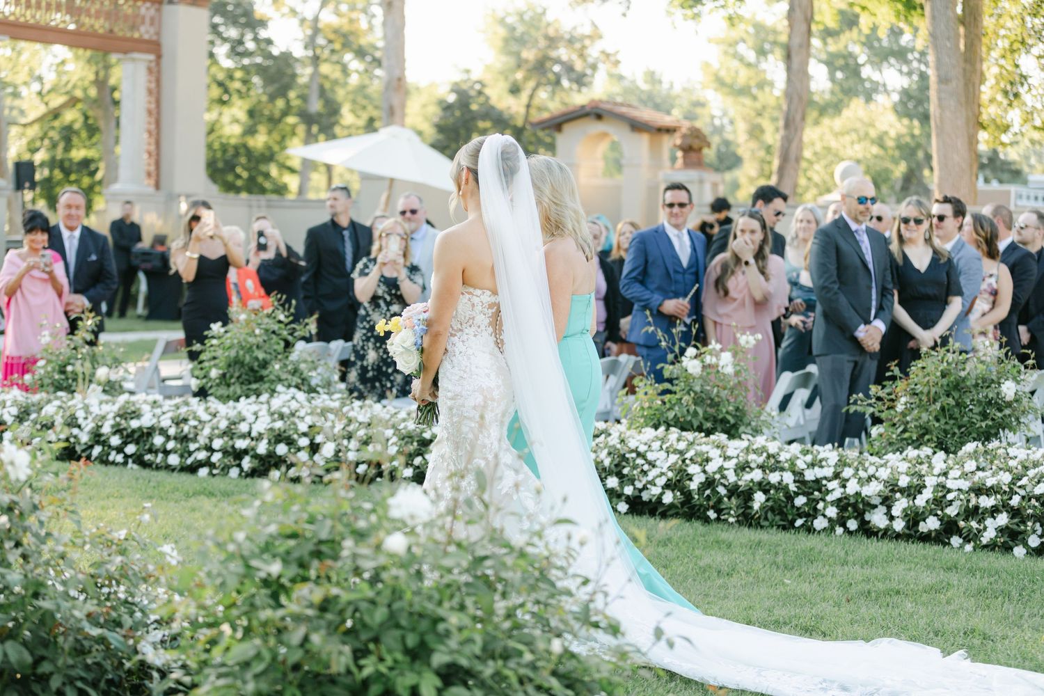 Armour House Wedding - Bride in white gown and veil walks down outdoor aisle lined with white flowers as wedding guests look on.