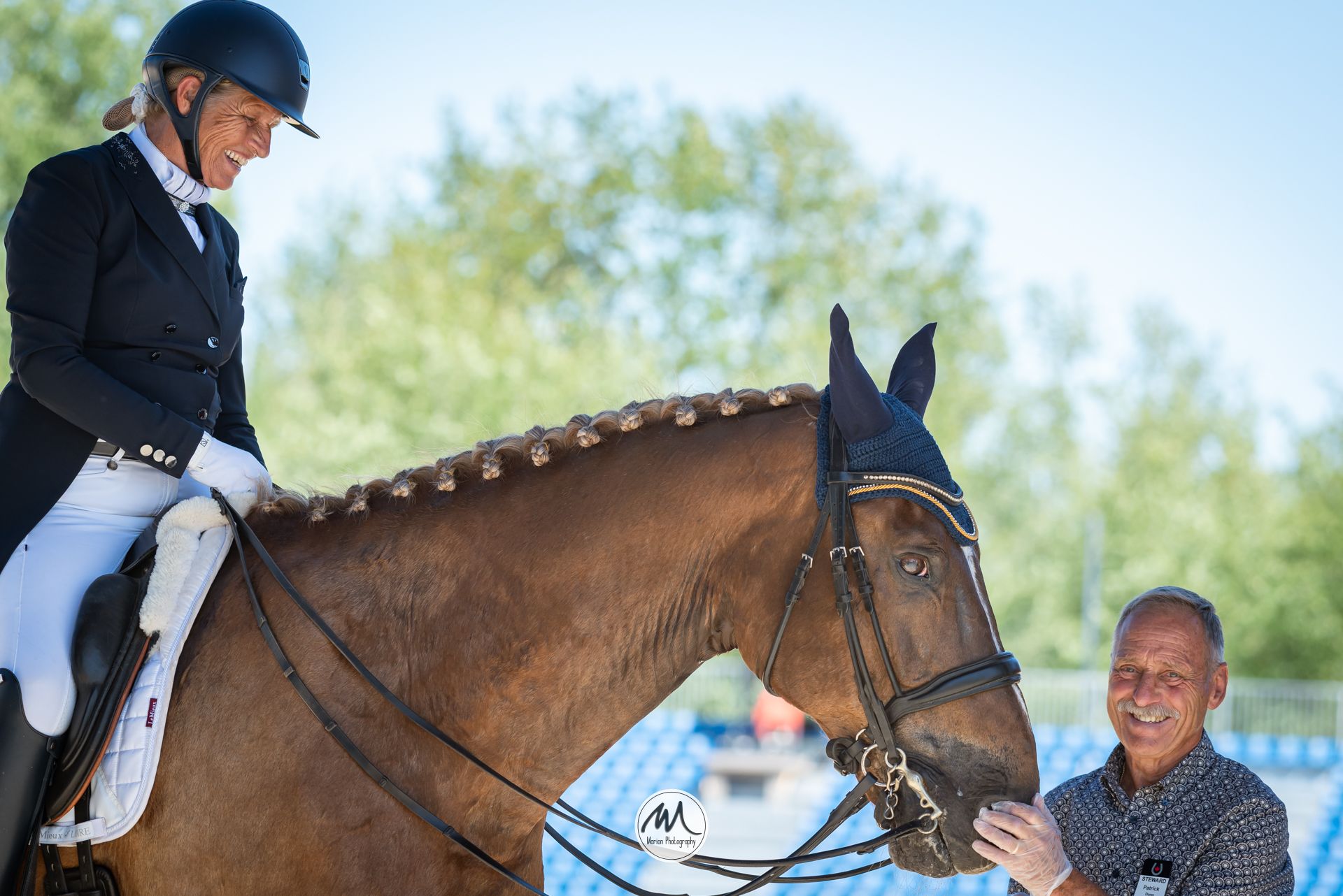 Equestrian rider in black jacket atop chestnut horse, with smiling man holding bridle at outdoor dressage competition.