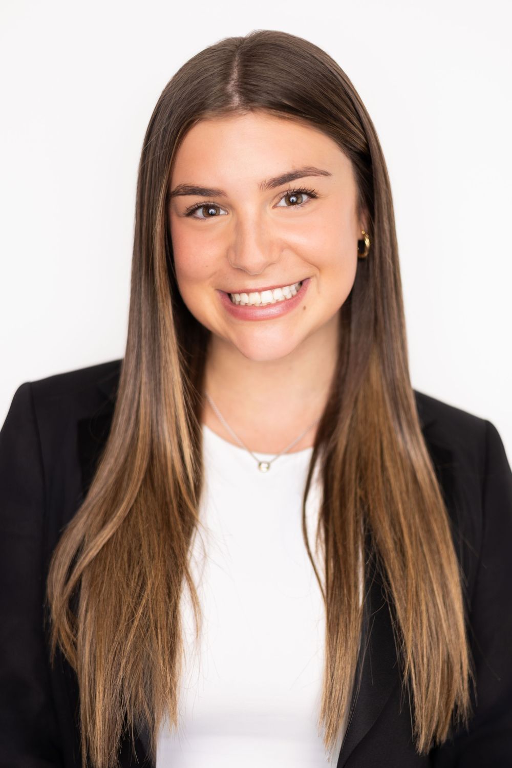 Young professional woman smiling in black blazer and white top, long brown hair, white background headshot.