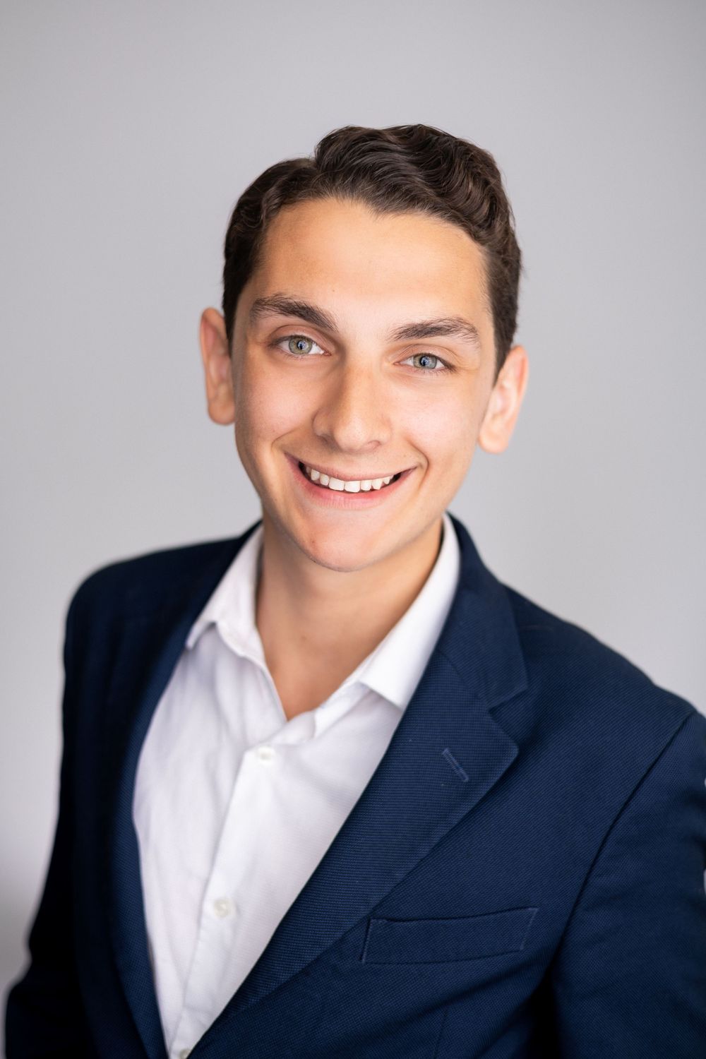 Young professional man smiling in navy blazer and white shirt against gray background.
