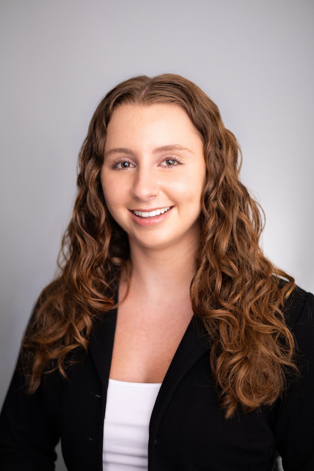 Young professional woman with curly auburn hair smiling confidently in a black blazer against a gray background.