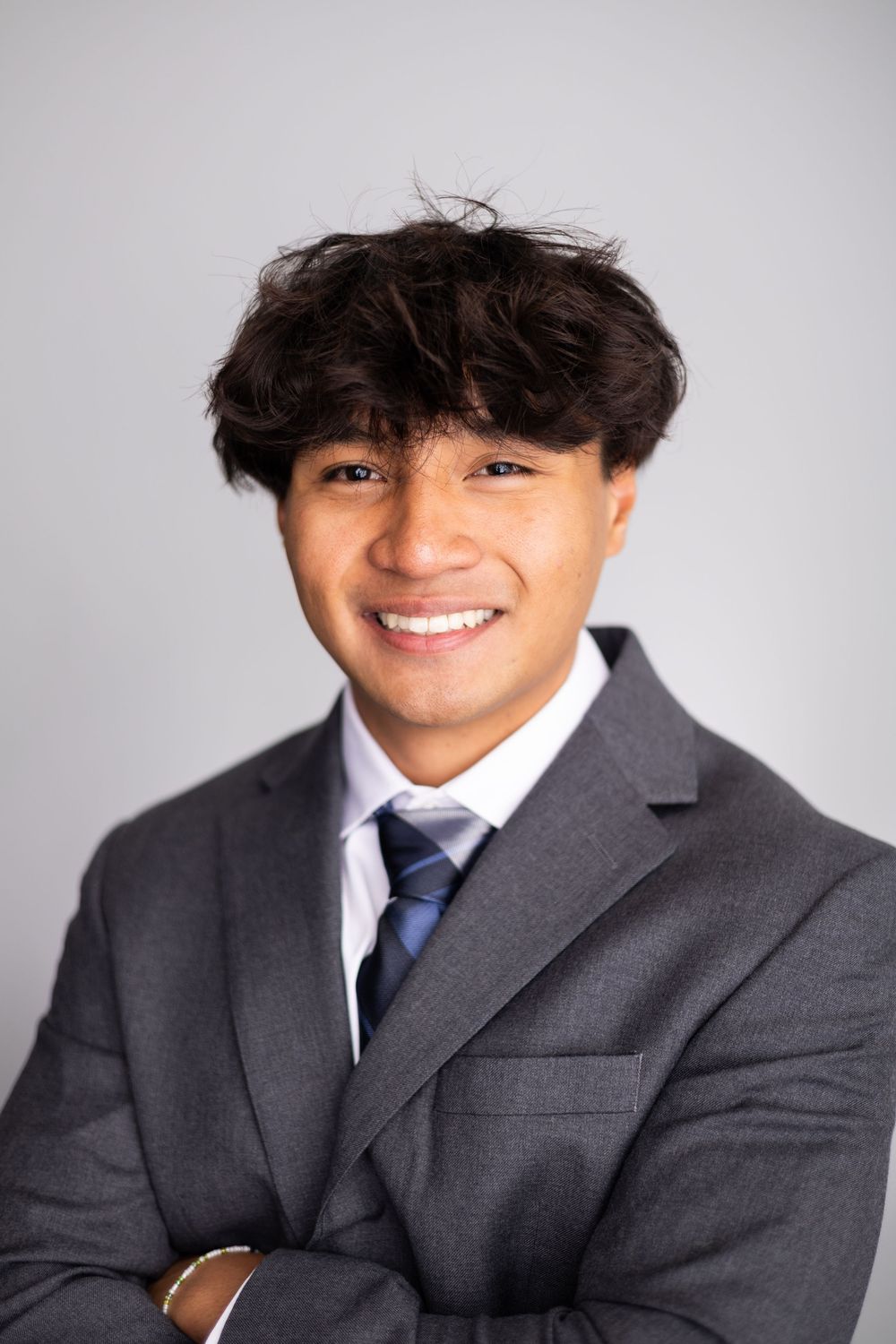 Young man in gray suit and blue tie smiling with arms crossed against gray background.
