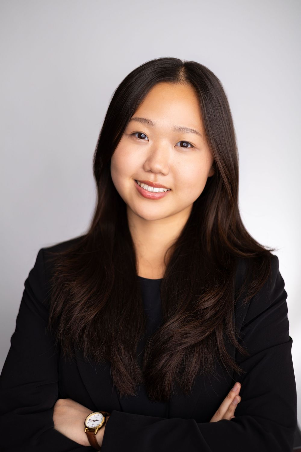 Professional headshot of a smiling young woman in a black blazer with arms crossed against a gray background.