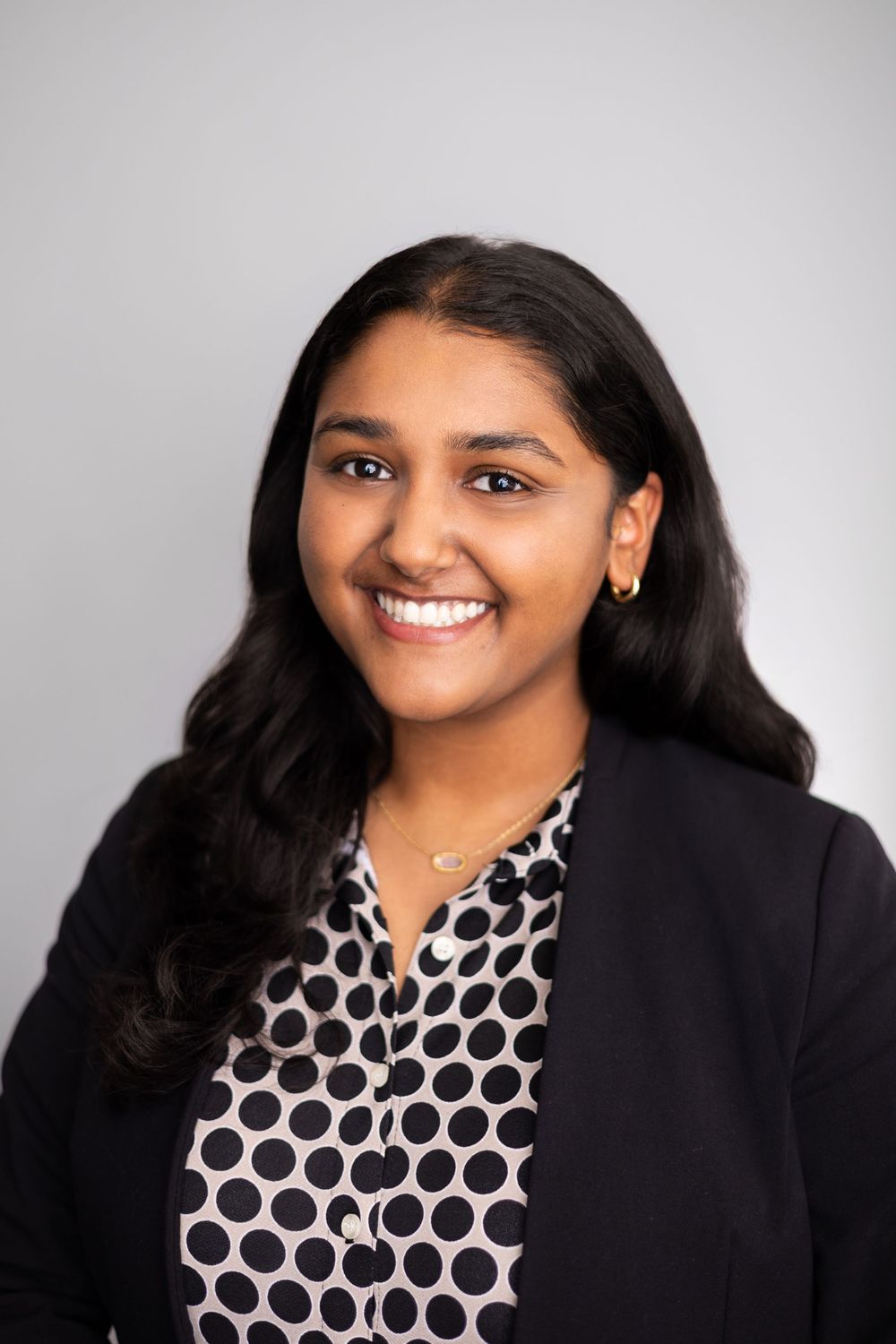 Professional headshot of a young woman in a black blazer and polka dot blouse, smiling confidently against a gray background.