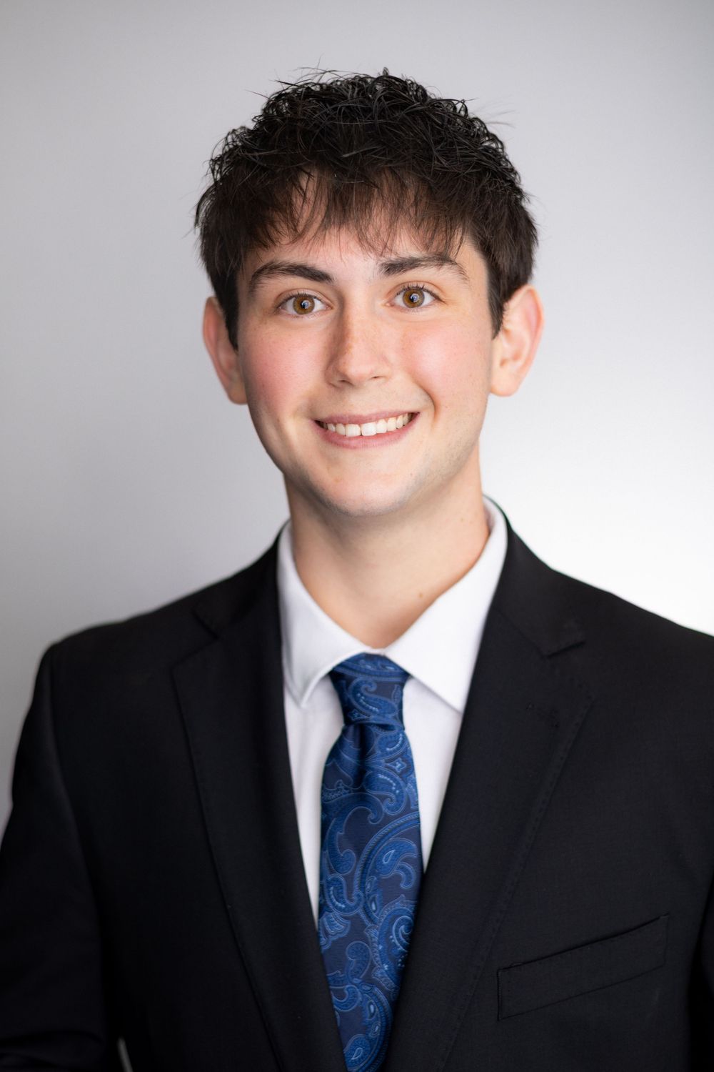Young professional man smiling in black suit and blue paisley tie, formal headshot photo.