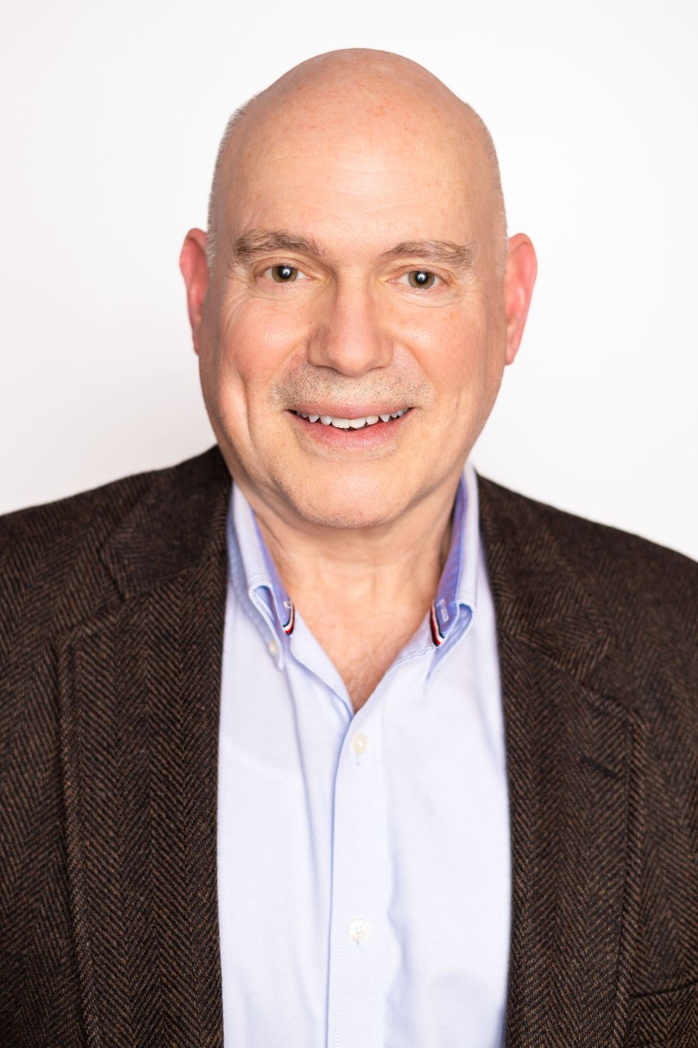 Smiling bald man in a brown herringbone blazer and light blue shirt poses for a professional headshot.