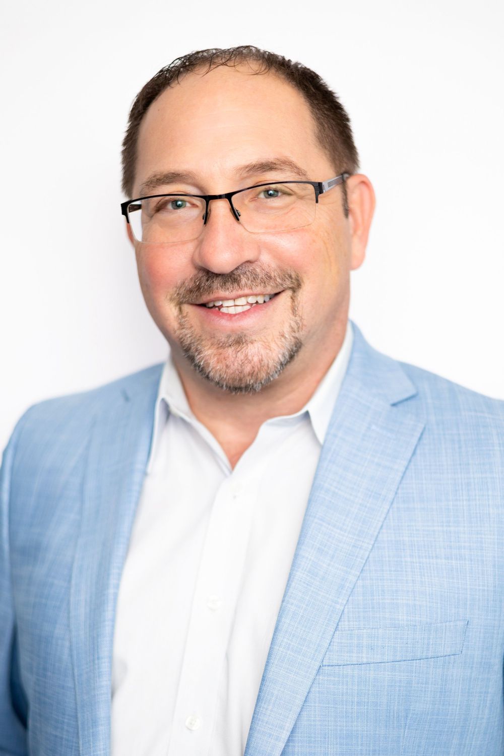 Smiling middle-aged man with glasses and stubble wearing a light blue blazer and white shirt against a white background.