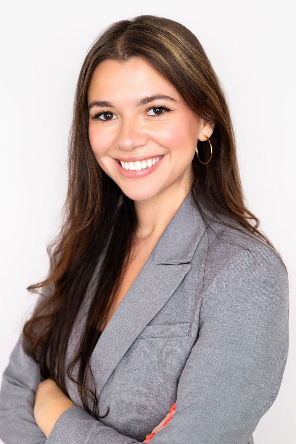 Young professional woman in gray blazer smiling confidently with arms crossed against white background.