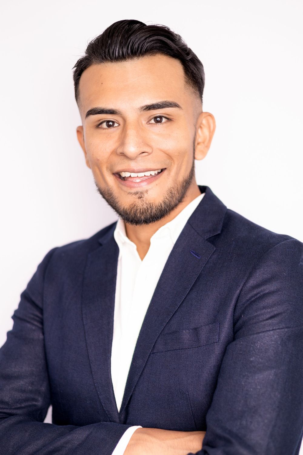 Smiling young businessman in navy blazer and white shirt with arms crossed against white background.