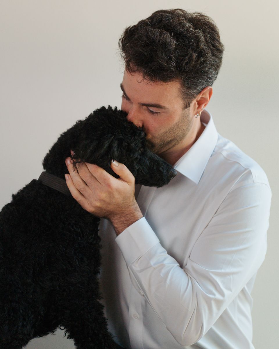A man in a white dress shirt affectionately kisses a fluffy black poodle dog against a light background. Dog photoshoot in Gables Hills