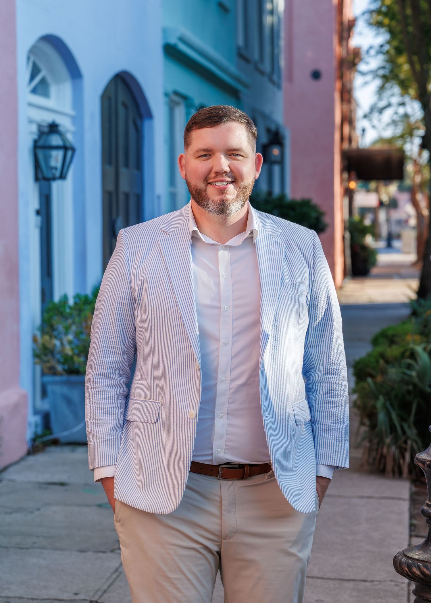 Man in light blue seersucker blazer smiling on colorful Charleston Rainbow Row street sidewalk.