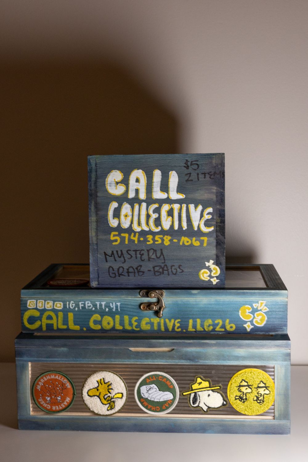 Handpainted blue wooden boxes displaying Call Collective phone number 574-358-1067, Peanuts characters, and mystery grab bags sign.