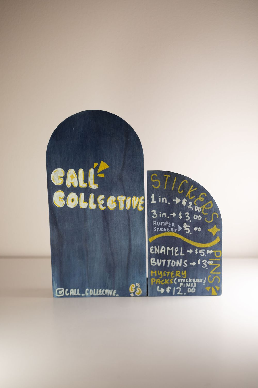 Dark blue wooden sign display for Call Collective showing sticker and enamel pin prices in yellow handwritten text.