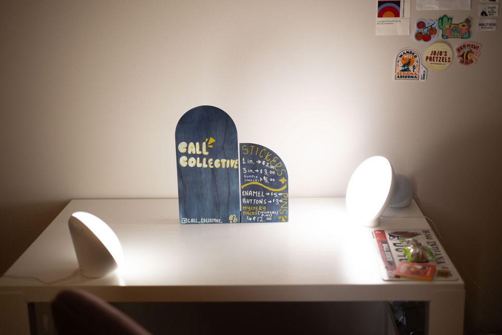 A desk with two white lamps flanking a blue arched sign reading Gallery Collective, with stickers on the wall behind.