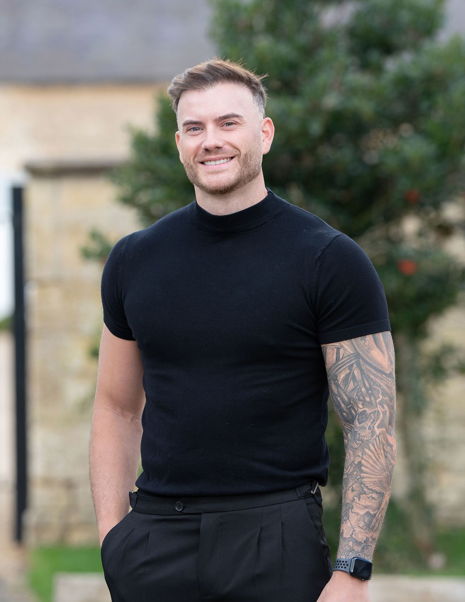 Smiling young man with tattoo sleeve wearing black turtleneck t-shirt and trousers, posing outdoors near green trees.
