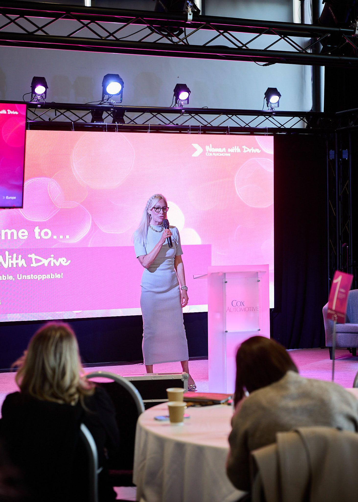 Woman speaker presenting at Women With Drive conference event with pink projection screen backdrop.