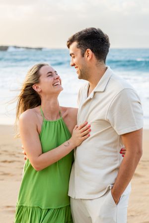 Happy couple laughing together on a sunny beach with ocean waves and blue sky behind.