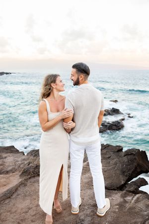 Couple in white outfits embracing on rocky shore with ocean waves at sunset in Hawaii.