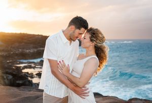 Romantic couple embracing at sunset on rocky Hawaii coastline with turquoise ocean waves.