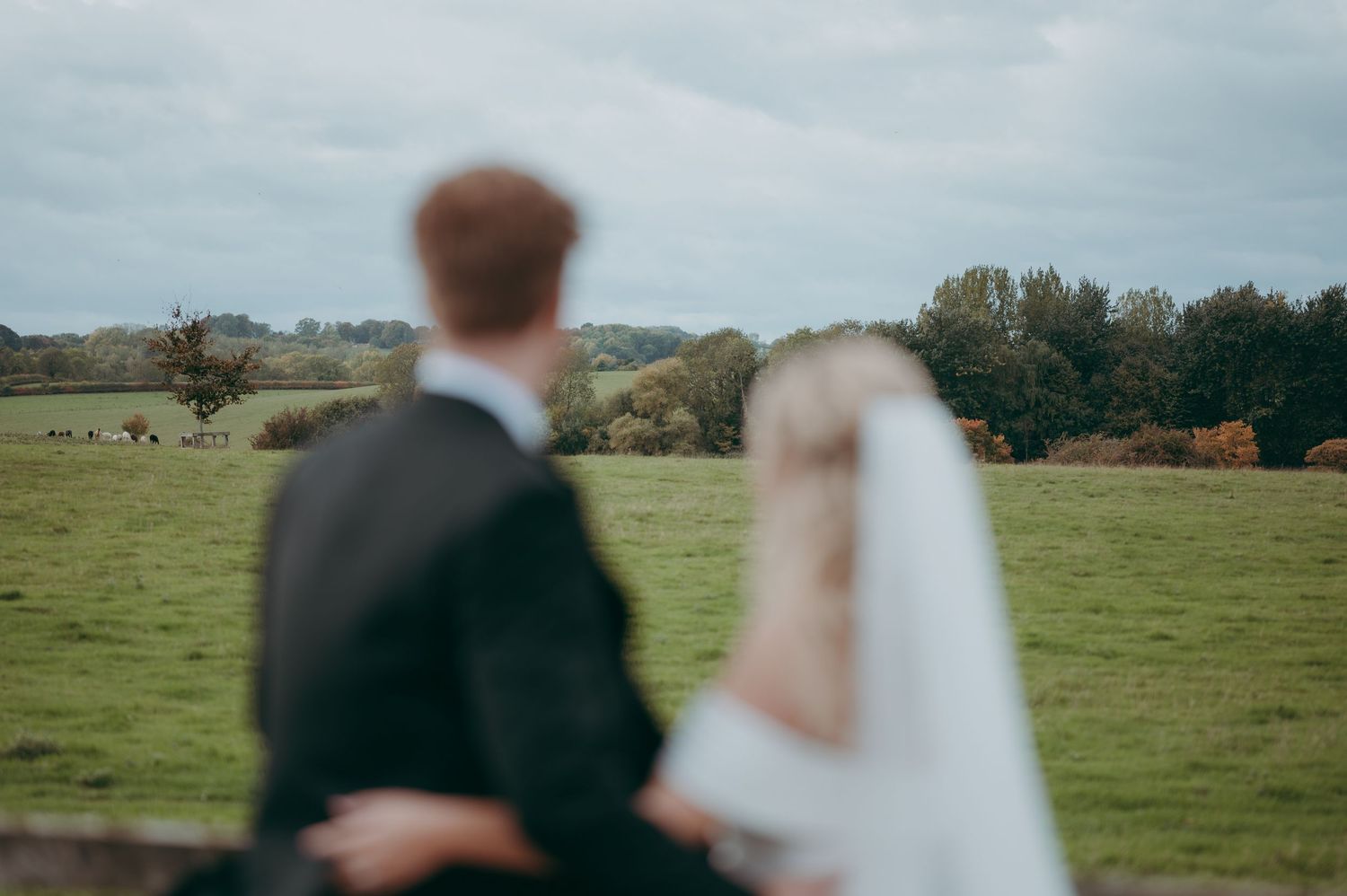 A bride and groom stand together overlooking a lush green countryside field on their wedding day.