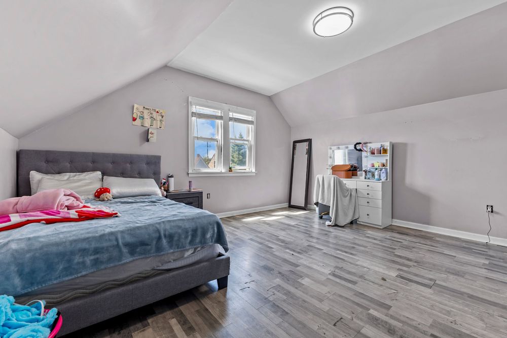Bright attic bedroom featuring vaulted ceilings, hardwood floors, a tufted headboard bed, and a window.