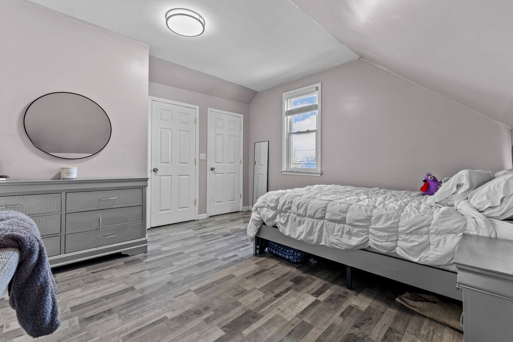 Cozy attic bedroom with sloped ceilings, gray dresser, round mirror, white door, and hardwood floors.