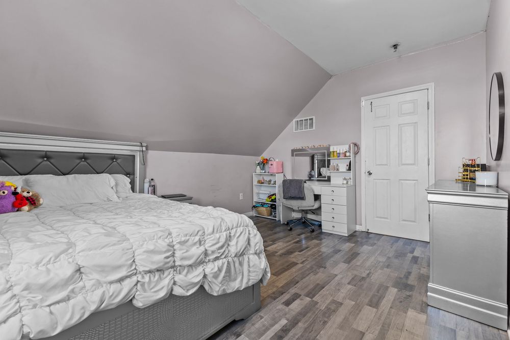 Attic bedroom with sloped ceiling, hardwood floors, white bed, desk area, and white door.