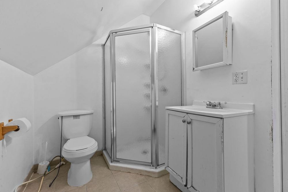 Small attic bathroom with sloped ceiling, corner shower, toilet, and white vanity with mirror cabinet.