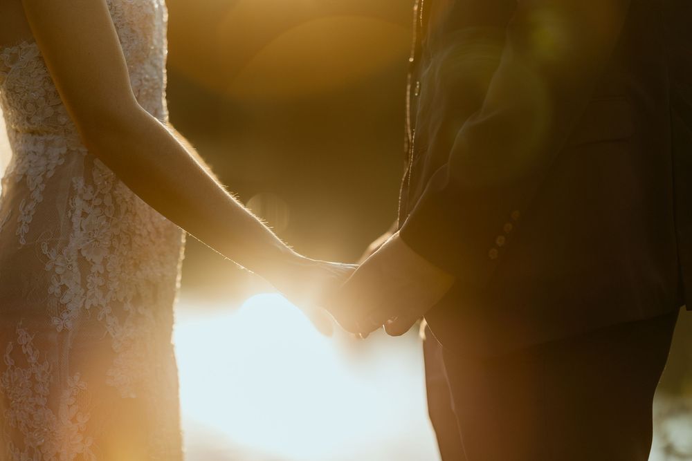 Bride and groom holding hands in warm golden sunset light, wedding dress and suit visible.