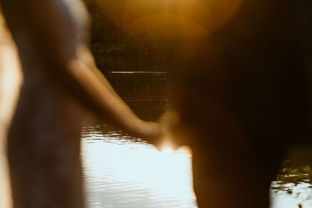 Couple holding hands at golden hour near a reflective lake, silhouetted in warm sunlight.