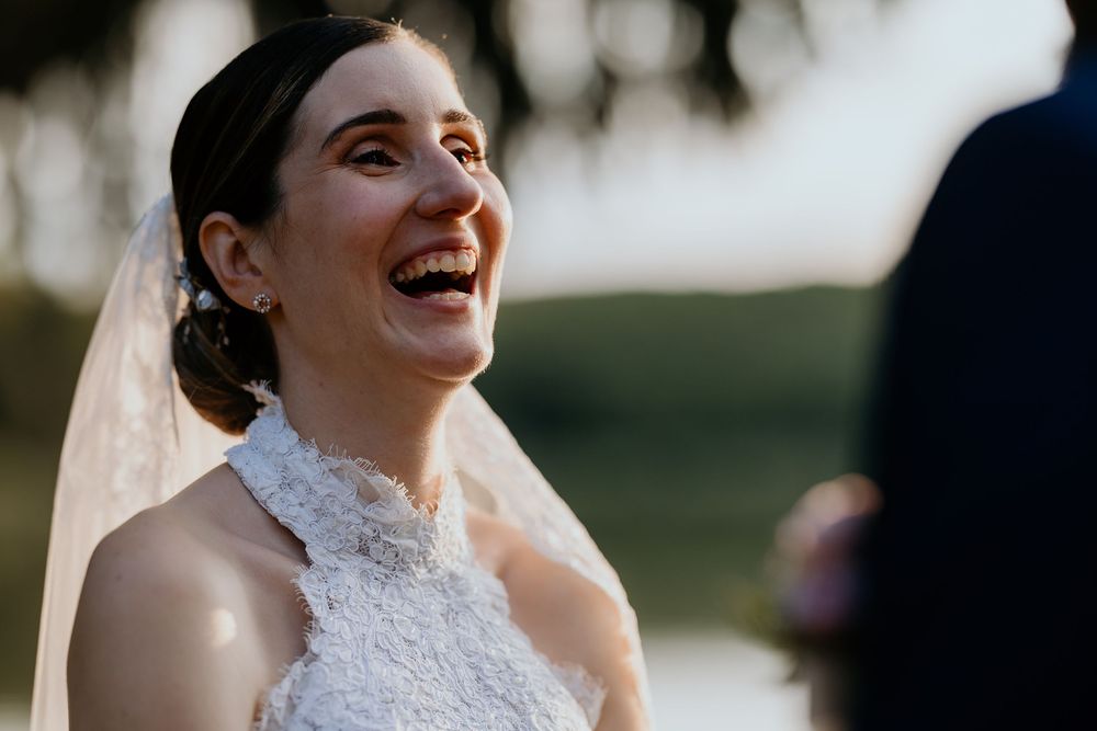 Bride laughing joyfully during outdoor wedding ceremony at golden hour near a lake.