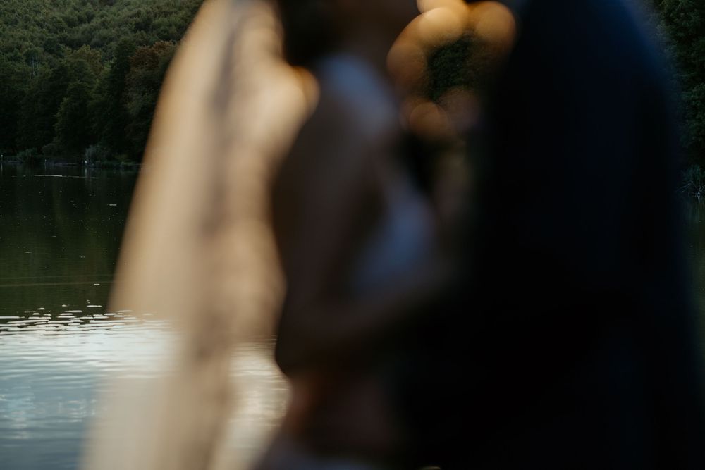 Artistic blurred shot of bride and groom near a lake with veil in foreground.