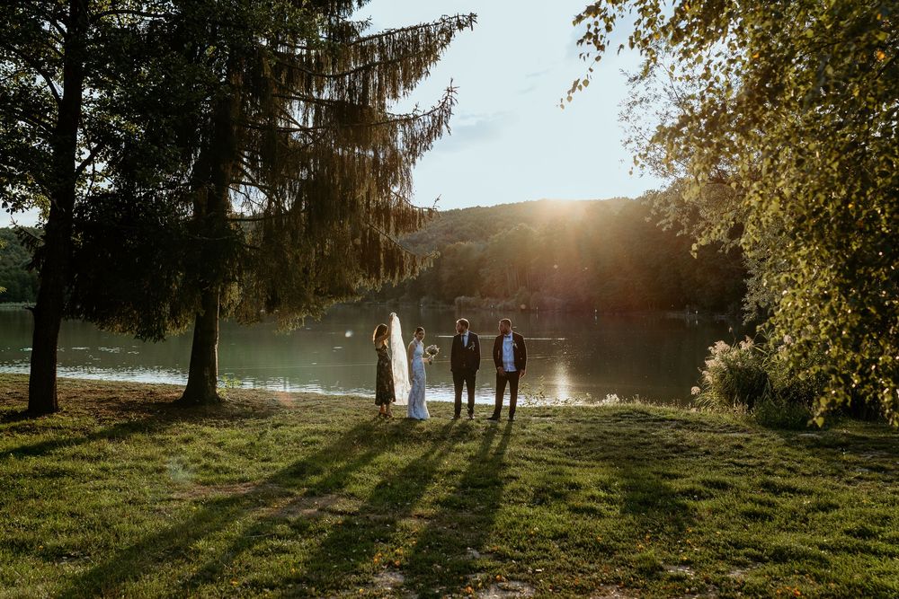 Wedding party standing by a sunlit lake surrounded by tall trees at golden hour.