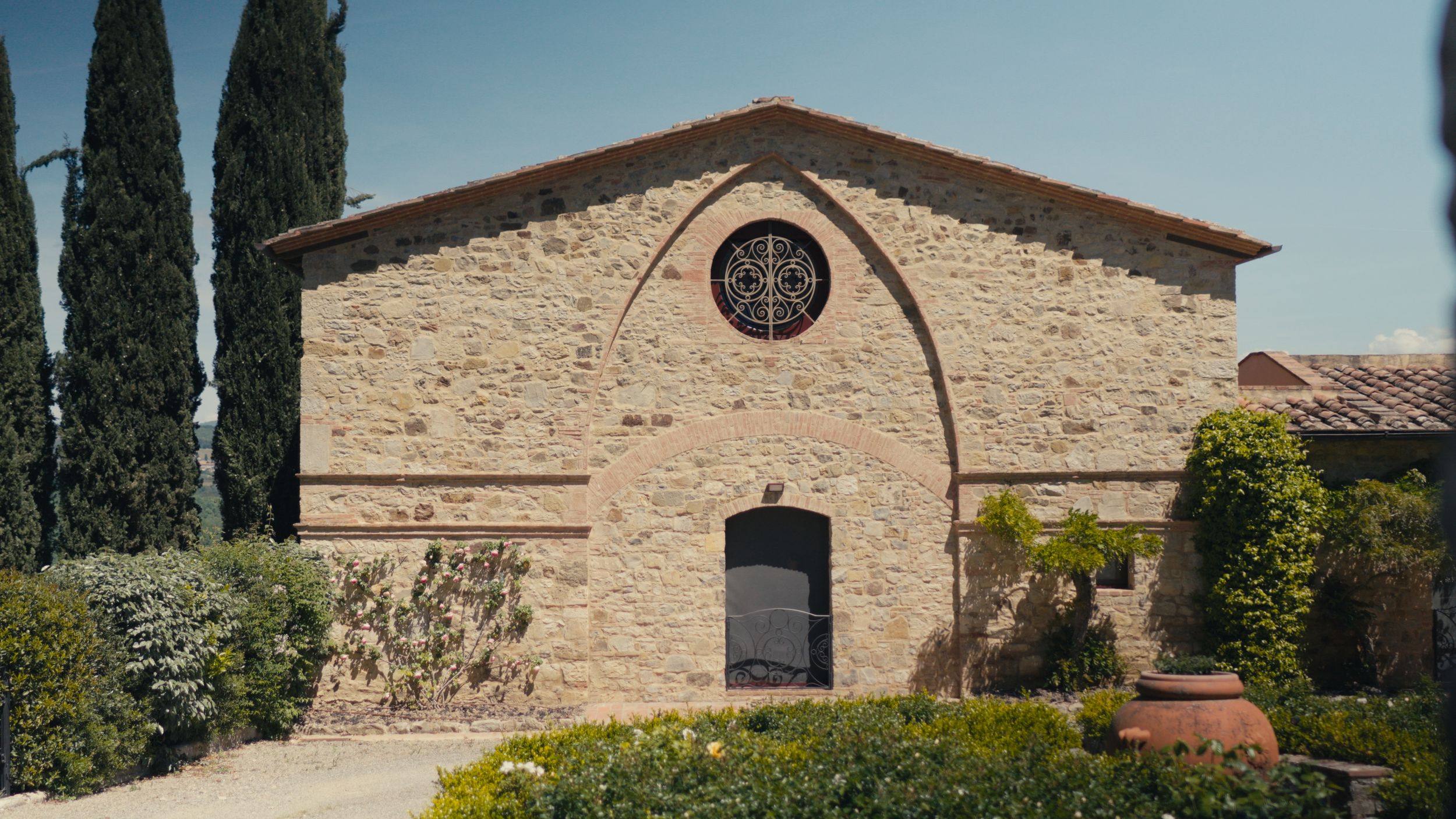 Stone Tuscan building with arched entrance, rose window, and cypress trees under a clear blue sky.