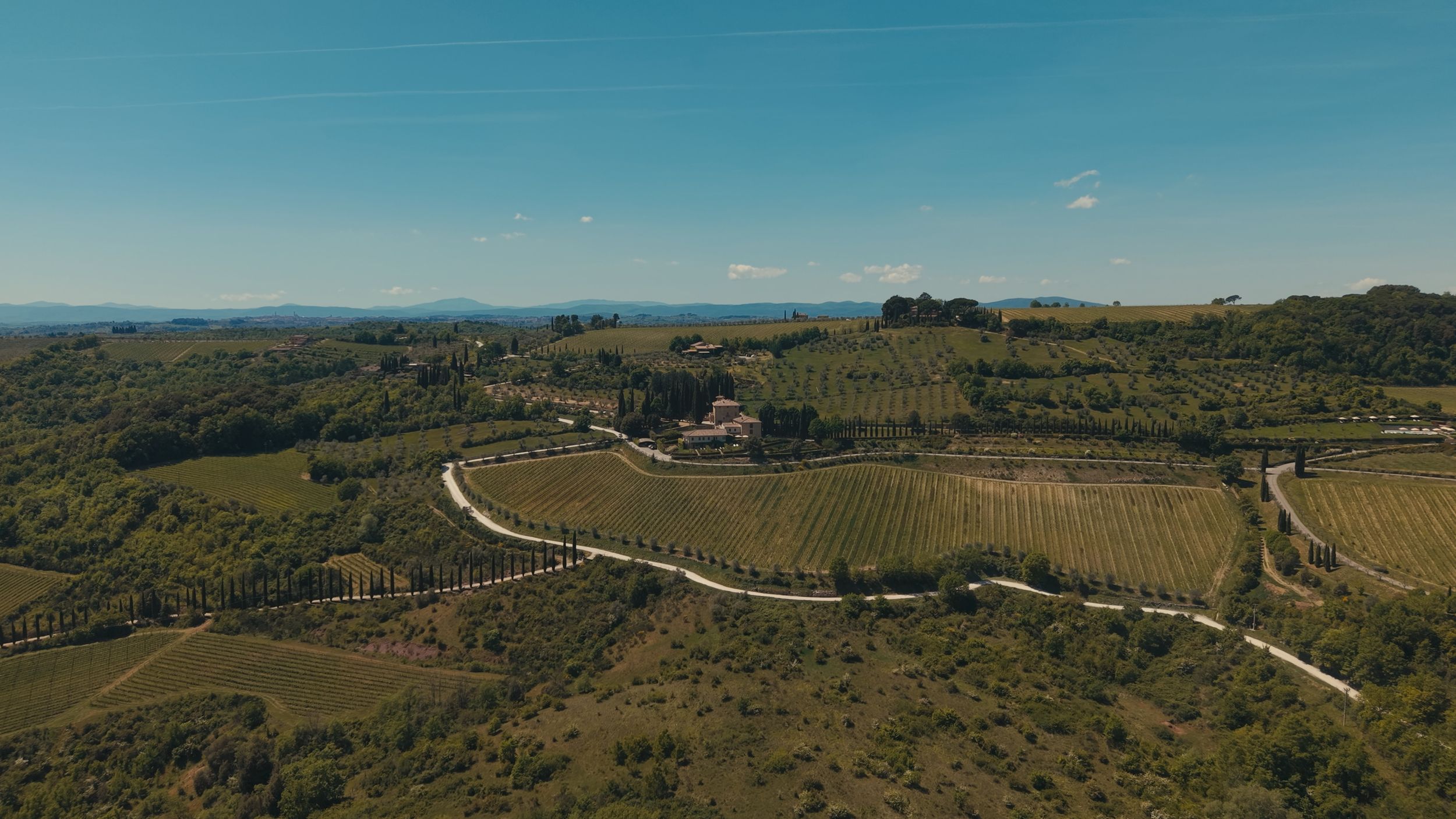 Aerial view of rolling Tuscan hills with vineyards, winding roads, and a farmhouse under a clear blue sky.